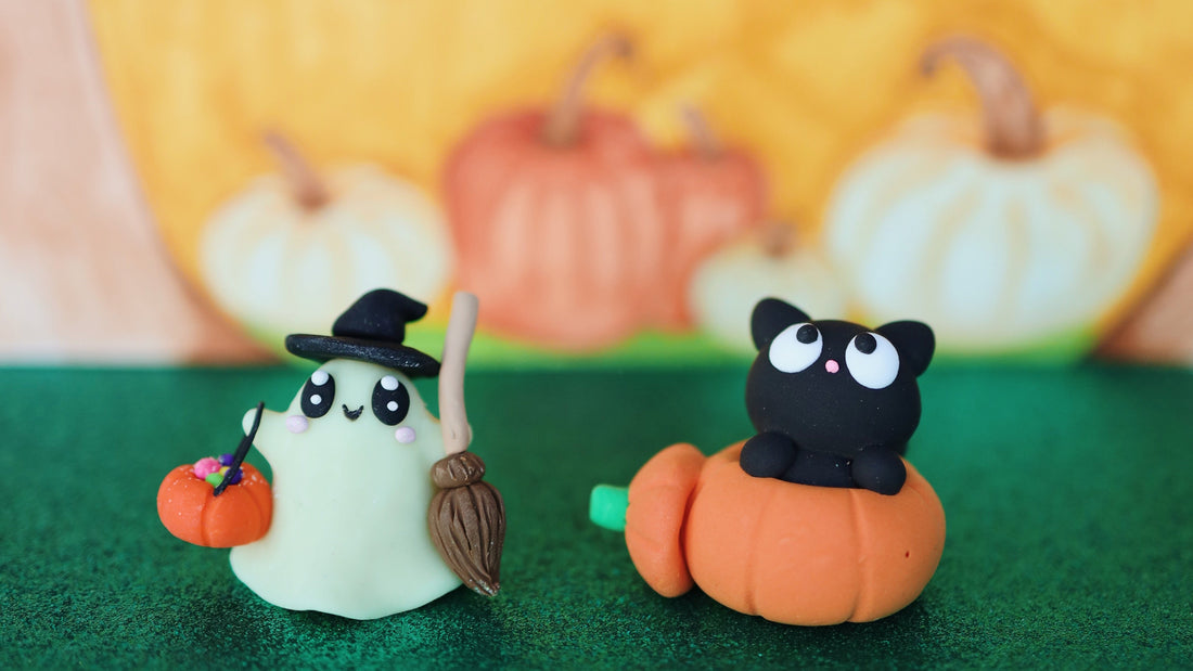 Two small Halloween-themed figurines on a green textured surface with a blurred pumpkin backdrop. One white ghost-like figure wears a black witch hat and holds a broom and a small orange pumpkin.
